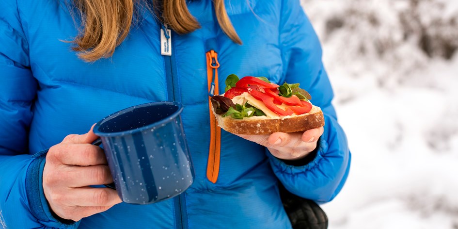 En person i vinterjacka håller i en mugg och en smörgås utomhus mot snöig bakgrund