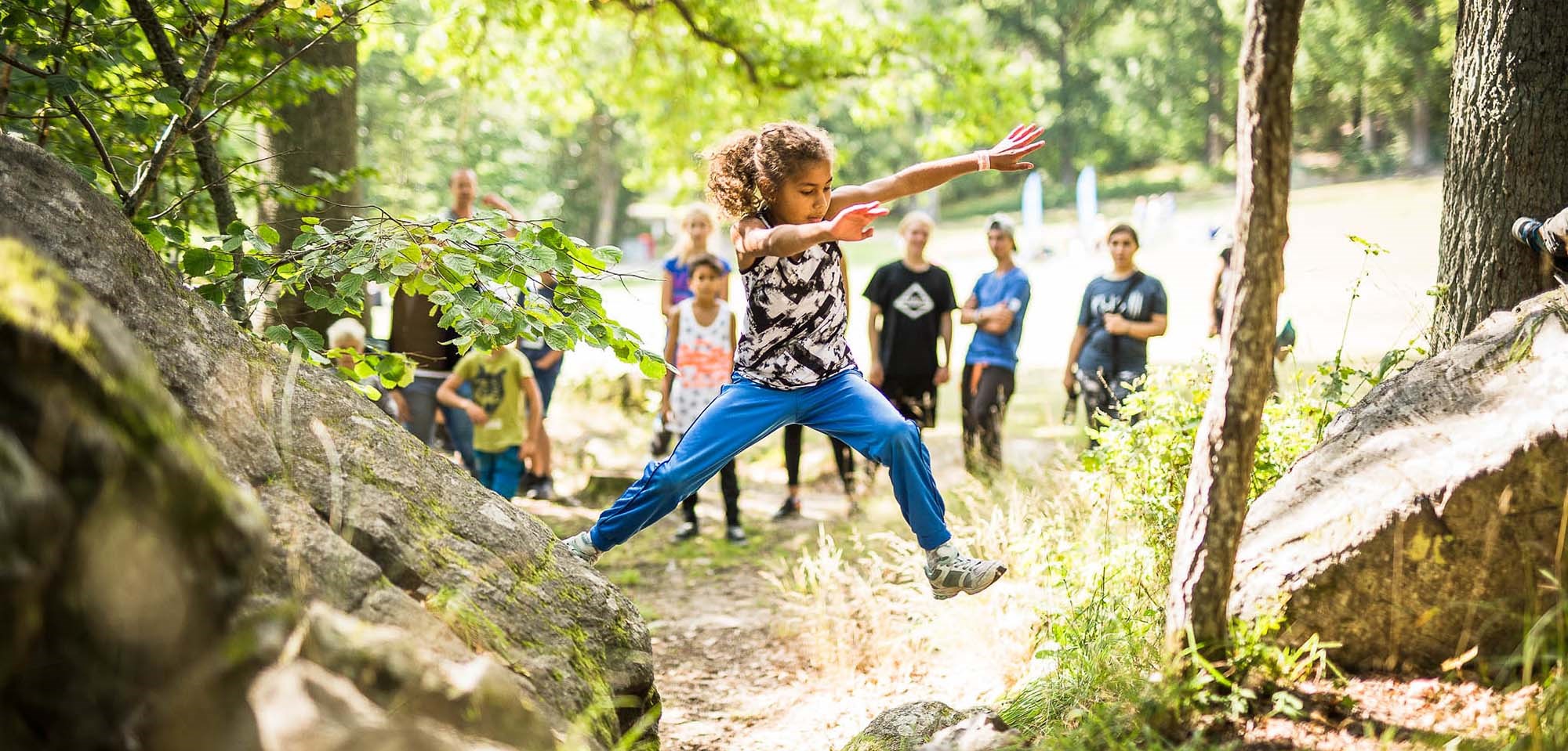 Barn hoppar mellan två stenar i skogen medan ledare och barngrupp ser på
