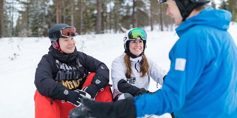 Två personer i skidskolan sitter i backen och lyssnar på sin skidlärare