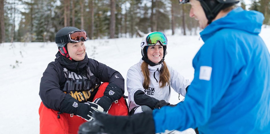 Två personer i skidskolan sitter i backen och lyssnar på sin skidlärare