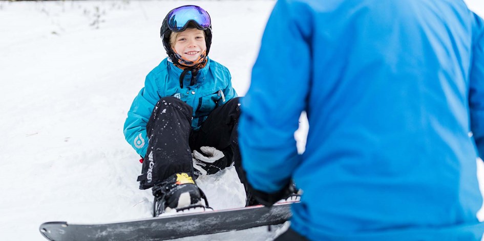 Barn med snowboard sitter vid backen tillsammans med ledare